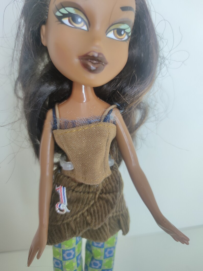 May include: A brown-skinned doll with long black hair wearing a brown corduroy skirt, a brown and gold corset, and green and blue checkered pants.