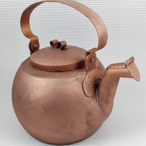 May include: A copper-coloured kettle with a rounded body, a curved handle, and a spout. The lid sits atop the kettle, and the handle arches over the top. The kettle is made of metal and has a vintage appearance.
