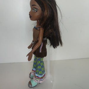 May include: A brown-haired doll with green eyes and brown eyeshadow wearing a brown corduroy top, blue and green patterned pants, and light blue shoes. The doll is standing on a clear plastic stand.