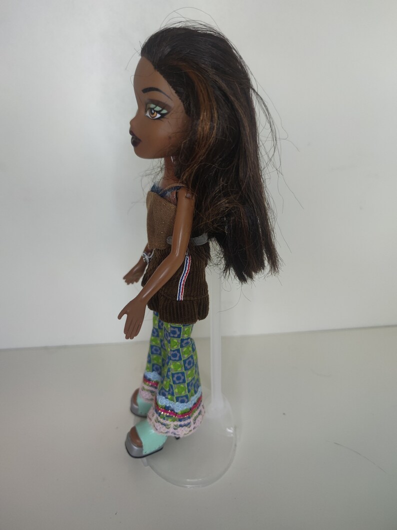 May include: A brown-haired doll with green eyes and brown eyeshadow wearing a brown corduroy top, blue and green patterned pants, and light blue shoes. The doll is standing on a clear plastic stand.