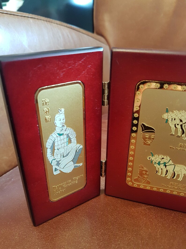 May include: A wooden display case with two hinged panels. The left panel features a gold-colored plaque with a white image of a warrior figure. The right panel features a gold-colored plaque with three white images of horses and a white image of a warrior's head. The text on the plaques reads "Terracotta Figure Of The Emperor's Army" and "The Emperor's Army".