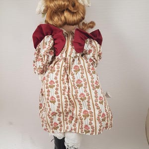 Puede incluir: Una mu&ntilde;eca de porcelana con cabello rojo, vestida con un vestido blanco con estampado floral y lazos rojos. La mu&ntilde;eca lleva botas negras y un borde de encaje blanco en el vestido.