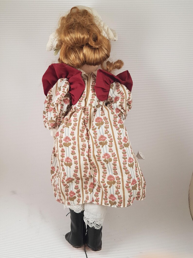 Puede incluir: Una mu&ntilde;eca de porcelana con cabello rojo, vestida con un vestido blanco con estampado floral y lazos rojos. La mu&ntilde;eca lleva botas negras y un borde de encaje blanco en el vestido.