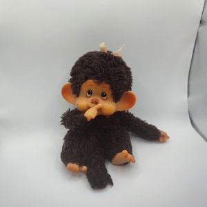 Puede incluir: Una muñeca Monchhichi vintage, con un cuerpo y cabeza de felpa marrón, y un mechón de pelo distintivo. La muñeca tiene grandes orejas naranjas, una cara clara con mejillas rosadas, y está posando con un dedo en la boca. Un juguete coleccionable clásico.