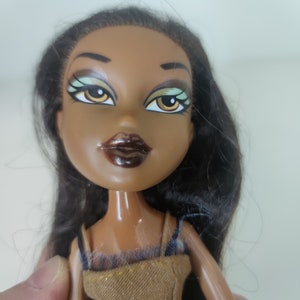 May include: A brown-skinned doll with long black hair, wearing a brown dress with a black lace overlay. The doll has green eyes with black eyeliner and brown lipstick.
