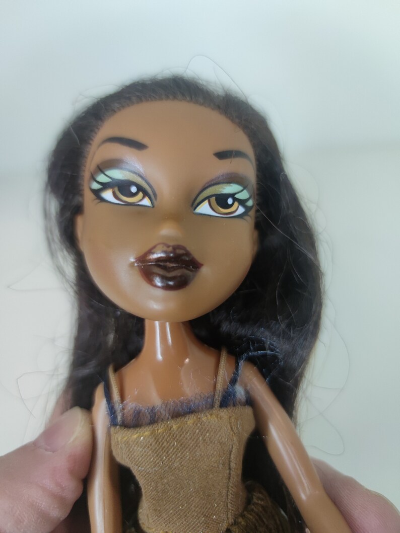 May include: A brown-skinned doll with long black hair, wearing a brown dress with a black lace overlay. The doll has green eyes with black eyeliner and brown lipstick.