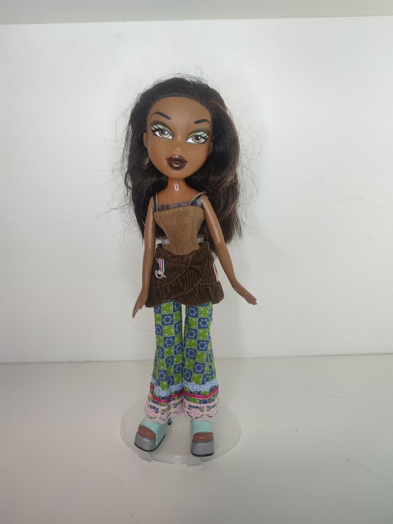 May include: A brown-skinned doll with long brown hair, wearing a brown corduroy top, green and blue patterned pants, and light blue platform shoes.