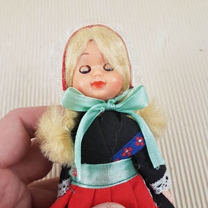 Puede incluir: Una peque&ntilde;a mu&ntilde;eca vestida con un vestido rojo, negro y blanco con una cinta verde. La mu&ntilde;eca tiene el pelo rubio y los ojos cerrados.