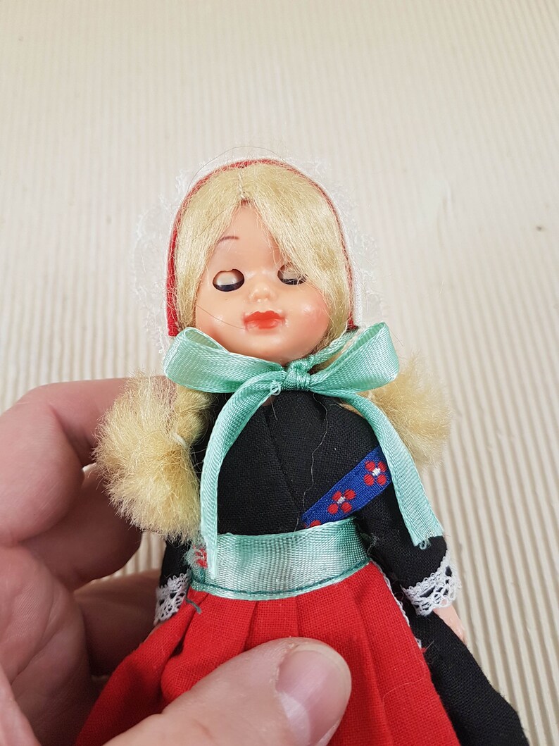 Puede incluir: Una peque&ntilde;a mu&ntilde;eca vestida con un vestido rojo, negro y blanco con una cinta verde. La mu&ntilde;eca tiene el pelo rubio y los ojos cerrados.