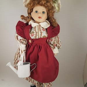 Puede incluir: Una mu&ntilde;eca de porcelana con pelo rojo, ojos azules y un sombrero blanco. Lleva un vestido rojo con un delantal blanco y un estampado floral. Lleva una regadera blanca.