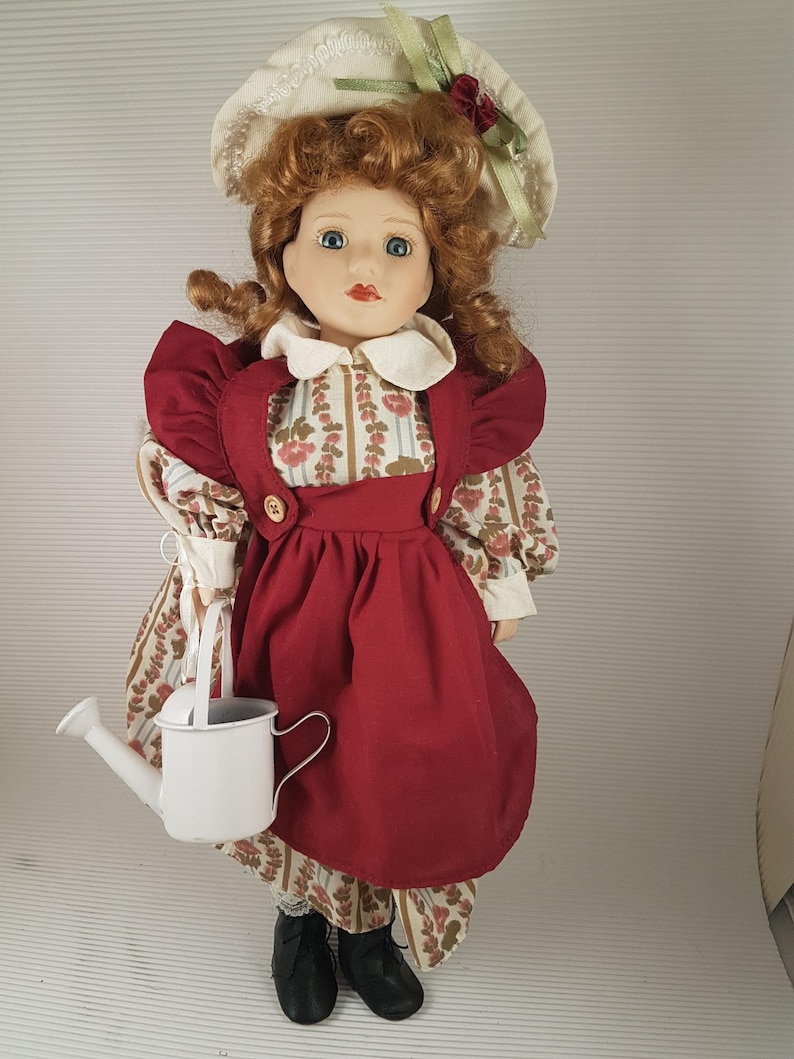 Puede incluir: Una mu&ntilde;eca de porcelana con pelo rojo, ojos azules y un sombrero blanco. Lleva un vestido rojo con un delantal blanco y un estampado floral. Lleva una regadera blanca.