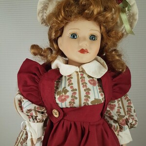Puede incluir: Una mu&ntilde;eca de porcelana con el pelo rojo rizado, que lleva un sombrero blanco con una cinta verde, un vestido rojo con estampado floral y un delantal blanco. La mu&ntilde;eca sostiene una regadera blanca.