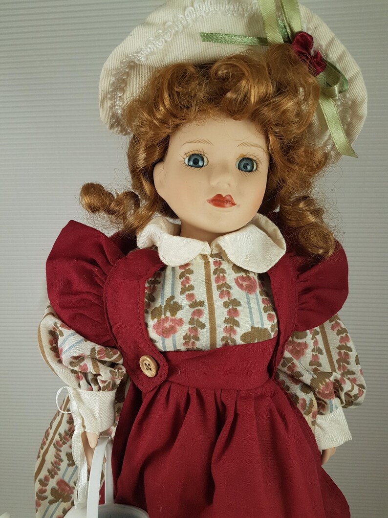 Puede incluir: Una mu&ntilde;eca de porcelana con el pelo rojo rizado, que lleva un sombrero blanco con una cinta verde, un vestido rojo con estampado floral y un delantal blanco. La mu&ntilde;eca sostiene una regadera blanca.