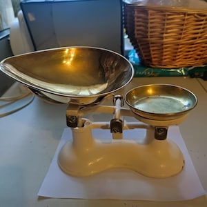 Puede incluir: Una antigua balanza blanca y de latón. La balanza tiene una gran bandeja ovalada de latón en un lado y una bandeja redonda más pequeña de latón en el otro. La base es blanca con un diseño curvo. La balanza probablemente se utiliza para medir el peso.