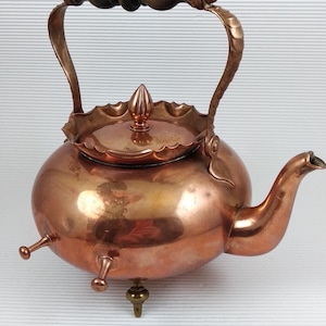 Può includere: Una teiera in rame con corpo arrotondato, beccuccio curvo e coperchio decorativo. Il manico è in legno e rame. La teiera ha tre piedini e un pomello decorativo sul coperchio. Un oggetto vintage per la cucina.