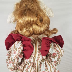 Puede incluir: Una mu&ntilde;eca de porcelana con un gorro blanco y un vestido rojo y blanco. La mu&ntilde;eca tiene el pelo largo, rizado y rubio.