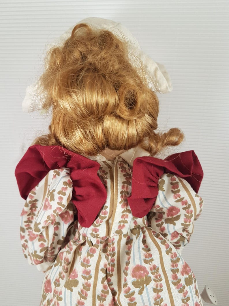 Puede incluir: Una mu&ntilde;eca de porcelana con un gorro blanco y un vestido rojo y blanco. La mu&ntilde;eca tiene el pelo largo, rizado y rubio.