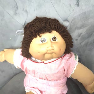 Puede incluir: Muñeca Cabbage Patch Kid vintage con cabello de hilo marrón y piel clara. La muñeca lleva un vestido a cuadros rosa y blanco con cuello y mangas blancos. La muñeca tiene ojos marrones y una cara pequeña y redonda.