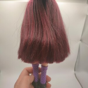 Pode incluir: Uma boneca com cabelo comprido, vermelho escuro e ra&iacute;zes pretas. A boneca est&aacute; usando meias de rede roxas e botas pretas. A boneca &eacute; segurada por uma m&atilde;o.