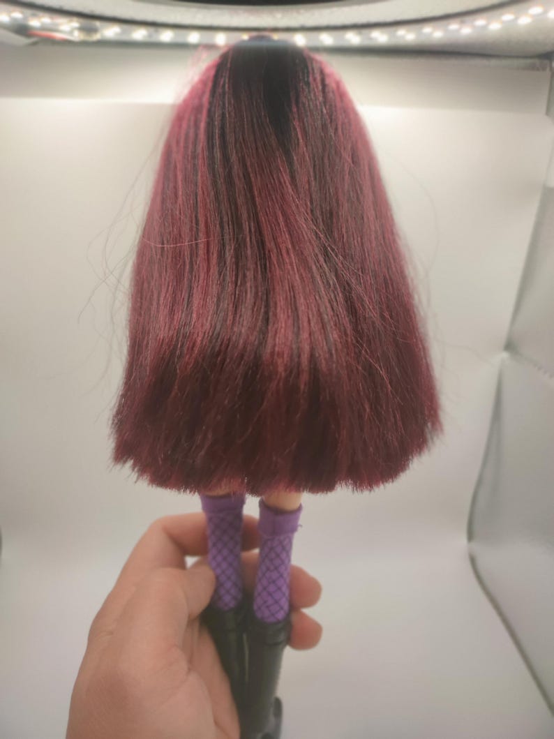 Pode incluir: Uma boneca com cabelo comprido, vermelho escuro e ra&iacute;zes pretas. A boneca est&aacute; usando meias de rede roxas e botas pretas. A boneca &eacute; segurada por uma m&atilde;o.