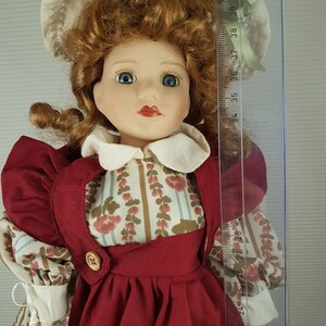 Puede incluir: Una mu&ntilde;eca de porcelana con el pelo rojo rizado, ojos azules y un vestido rojo con estampado floral. Lleva un sombrero blanco con ribete verde y sostiene una cesta blanca.
