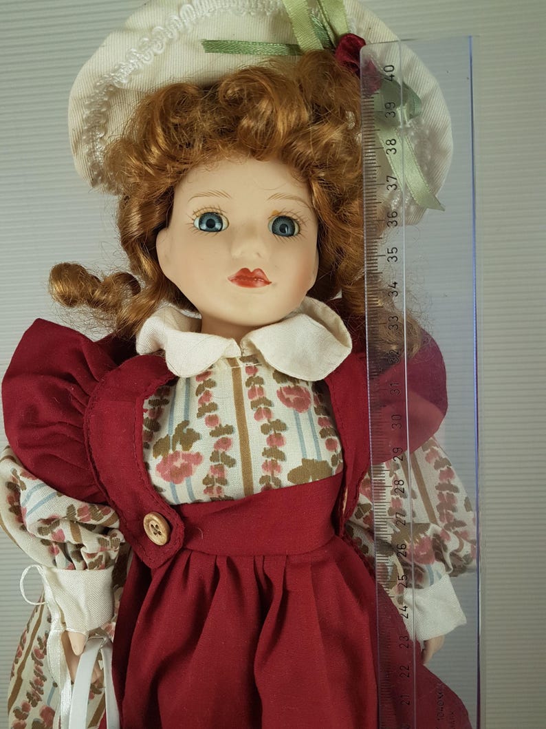 Puede incluir: Una mu&ntilde;eca de porcelana con el pelo rojo rizado, ojos azules y un vestido rojo con estampado floral. Lleva un sombrero blanco con ribete verde y sostiene una cesta blanca.