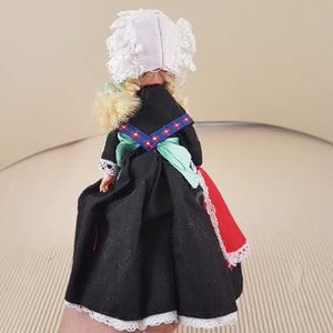 Puede incluir: Una peque&ntilde;a mu&ntilde;eca vestida con un vestido negro con ribetes de encaje blanco, una faja verde y un delantal rojo y blanco. La mu&ntilde;eca lleva una cofia blanca con ribetes de encaje y tiene el pelo rubio.