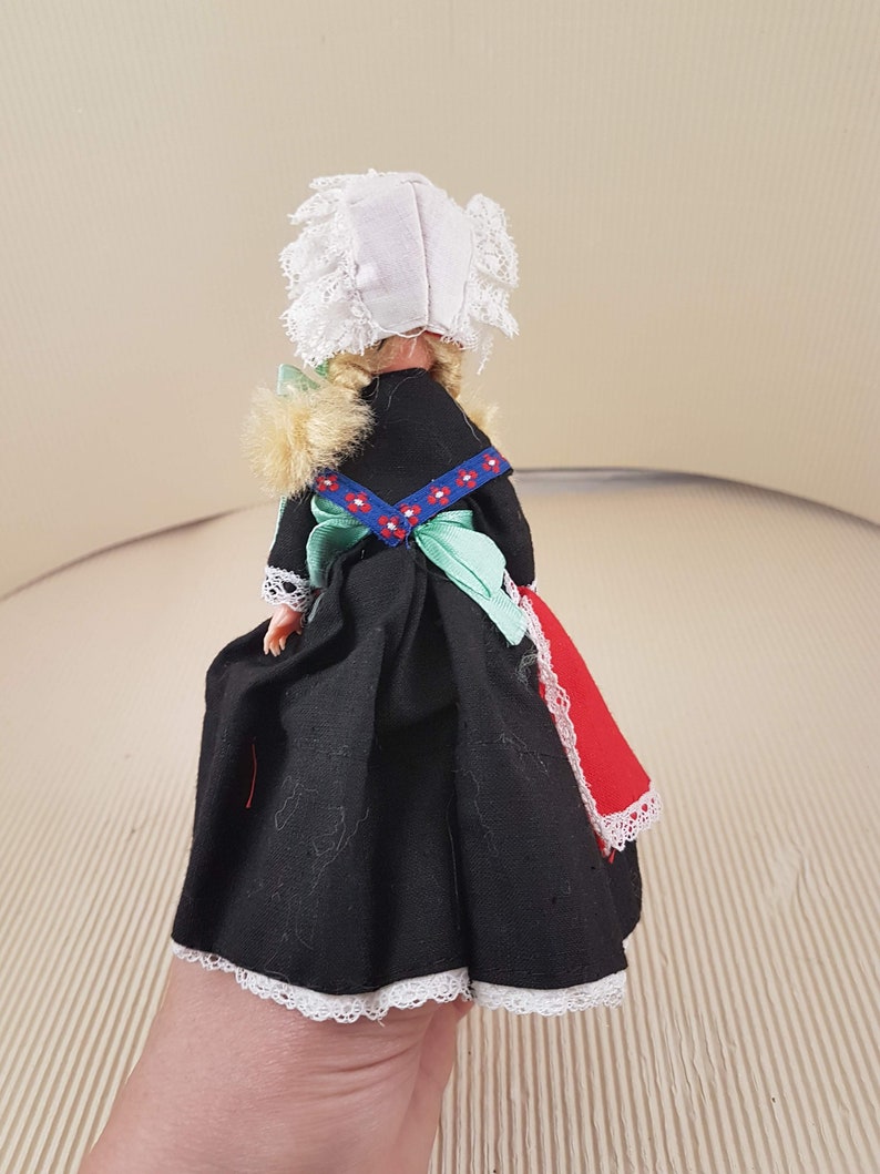 Puede incluir: Una peque&ntilde;a mu&ntilde;eca vestida con un vestido negro con ribetes de encaje blanco, una faja verde y un delantal rojo y blanco. La mu&ntilde;eca lleva una cofia blanca con ribetes de encaje y tiene el pelo rubio.