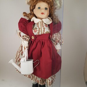Puede incluir: Una mu&ntilde;eca de porcelana que lleva un vestido rojo con un delantal blanco y un sombrero blanco con ribete verde. La mu&ntilde;eca sostiene una regadera blanca y est&aacute; de pie sobre una superficie blanca.