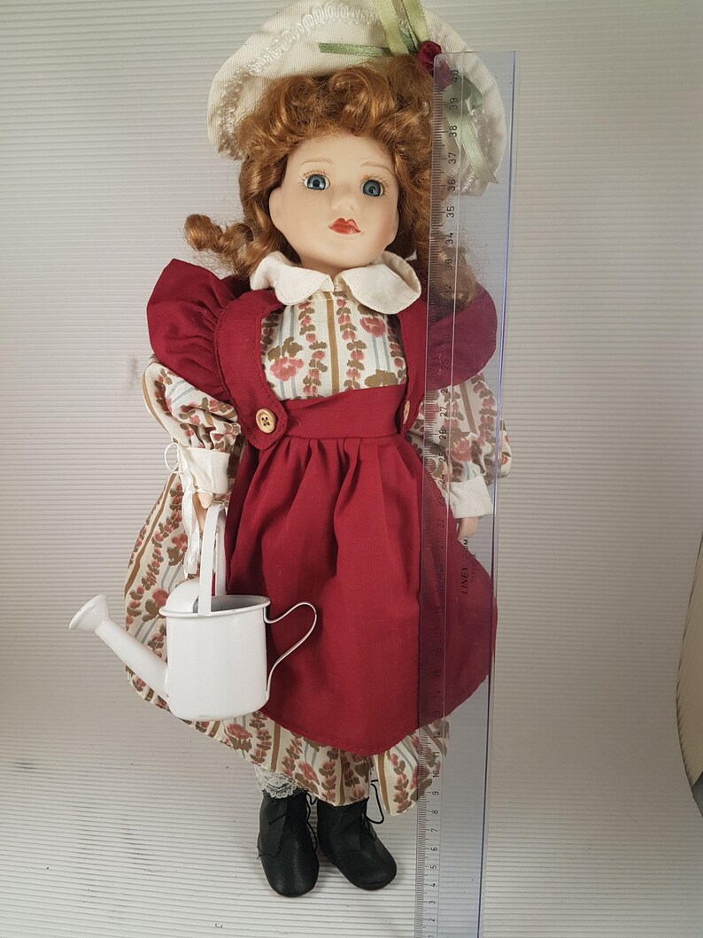 Puede incluir: Una mu&ntilde;eca de porcelana que lleva un vestido rojo con un delantal blanco y un sombrero blanco con ribete verde. La mu&ntilde;eca sostiene una regadera blanca y est&aacute; de pie sobre una superficie blanca.