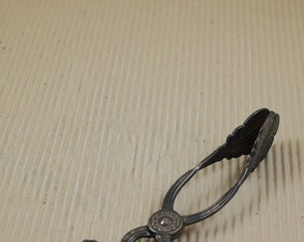 May include: Silver-toned serving tongs with an ornate design. The tongs are open and lying on a light brown surface.