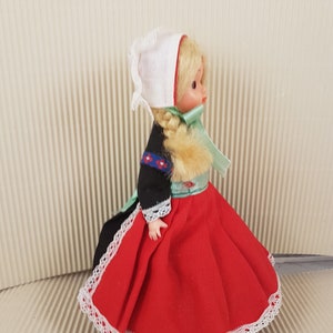 Puede incluir: Una peque&ntilde;a mu&ntilde;eca vestida con un traje tradicional holand&eacute;s. La mu&ntilde;eca tiene el pelo rubio, un gorro blanco, un corpi&ntilde;o negro, una falda roja con encaje blanco y una cinta verde.