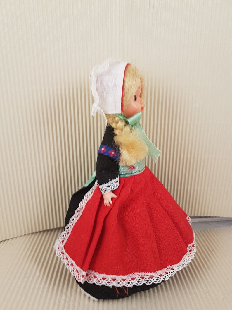Puede incluir: Una peque&ntilde;a mu&ntilde;eca vestida con un traje tradicional holand&eacute;s. La mu&ntilde;eca tiene el pelo rubio, un gorro blanco, un corpi&ntilde;o negro, una falda roja con encaje blanco y una cinta verde.