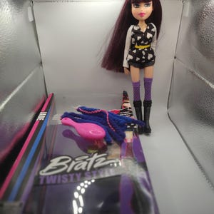 Pode incluir: Boneca Bratz Twisty Style com cabelo longo e escuro, usando um top preto e branco com estampa de cora&ccedil;&atilde;o, shorts e botas. Acess&oacute;rios incluem uma escova rosa e extens&otilde;es de cabelo azuis. A boneca est&aacute; ao lado da embalagem.