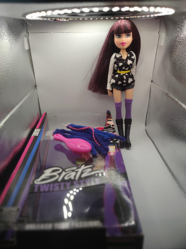 Pode incluir: Boneca Bratz Twisty Style com cabelo longo e escuro, usando um top preto e branco com estampa de cora&ccedil;&atilde;o, shorts e botas. Acess&oacute;rios incluem uma escova rosa e extens&otilde;es de cabelo azuis. A boneca est&aacute; ao lado da embalagem.