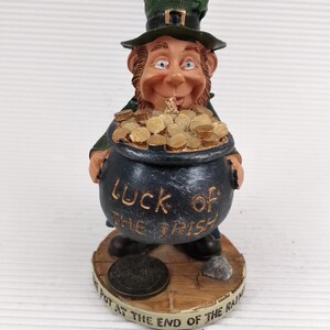 May include: A figurine of a leprechaun holding a pot of gold. The leprechaun wears a green hat and coat. The pot is black with the words "LUCK OF THE IRISH" on it. The base reads "THE POT AT THE END OF THE RAINBOW".