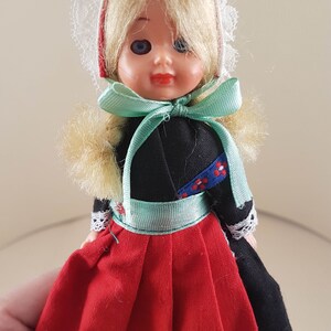 Puede incluir: Una peque&ntilde;a mu&ntilde;eca vestida con un traje tradicional holand&eacute;s. La mu&ntilde;eca tiene el pelo rubio, los ojos azules y un vestido rojo y negro con un borde de encaje blanco. La mu&ntilde;eca lleva una cofia de encaje blanco con una cinta roja.