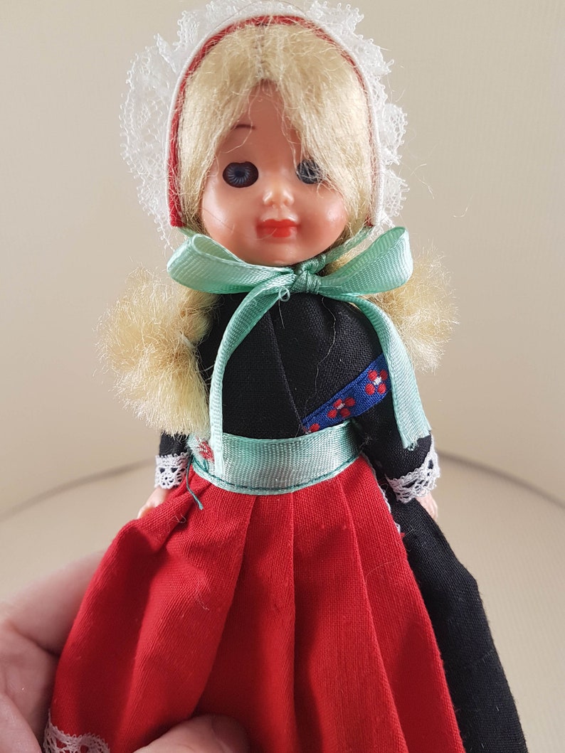 Puede incluir: Una peque&ntilde;a mu&ntilde;eca vestida con un traje tradicional holand&eacute;s. La mu&ntilde;eca tiene el pelo rubio, los ojos azules y un vestido rojo y negro con un borde de encaje blanco. La mu&ntilde;eca lleva una cofia de encaje blanco con una cinta roja.