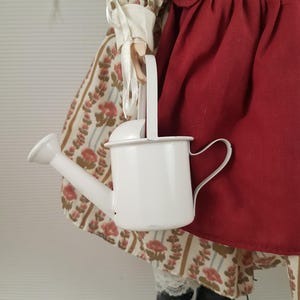 Puede incluir: Una mu&ntilde;eca de porcelana con un vestido rojo y blanco y sosteniendo una regadera blanca.