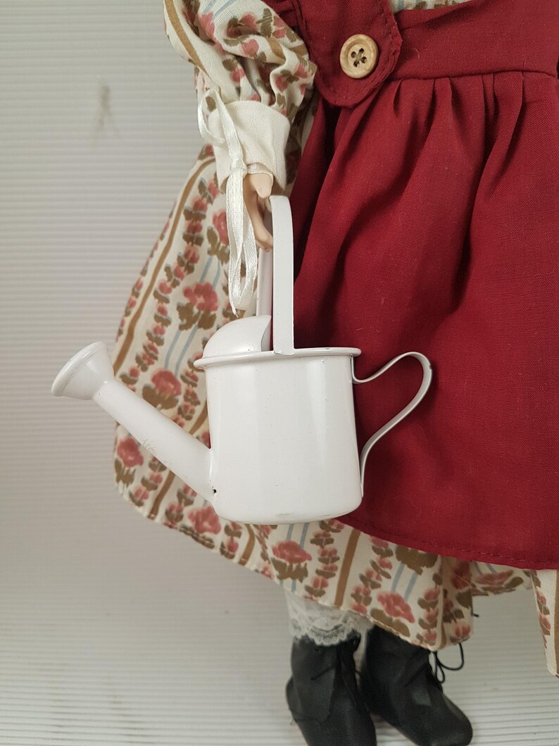 Puede incluir: Una mu&ntilde;eca de porcelana con un vestido rojo y blanco y sosteniendo una regadera blanca.
