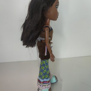 May include: A brown-haired doll with brown skin and green eyes is wearing a brown corduroy skirt, a blue and green patterned shirt, and blue and green shoes. The doll is standing on a clear plastic stand.