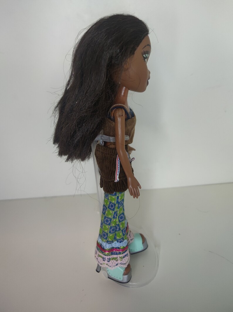 May include: A brown-haired doll with brown skin and green eyes is wearing a brown corduroy skirt, a blue and green patterned shirt, and blue and green shoes. The doll is standing on a clear plastic stand.