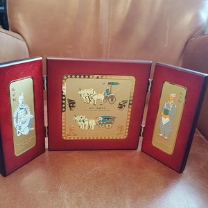 May include: A three-panel wooden display with gold-toned plaques depicting scenes from the Terracotta Army. The center panel shows a chariot pulled by four horses, while the side panels feature figures of warriors. The plaques are surrounded by a decorative border.