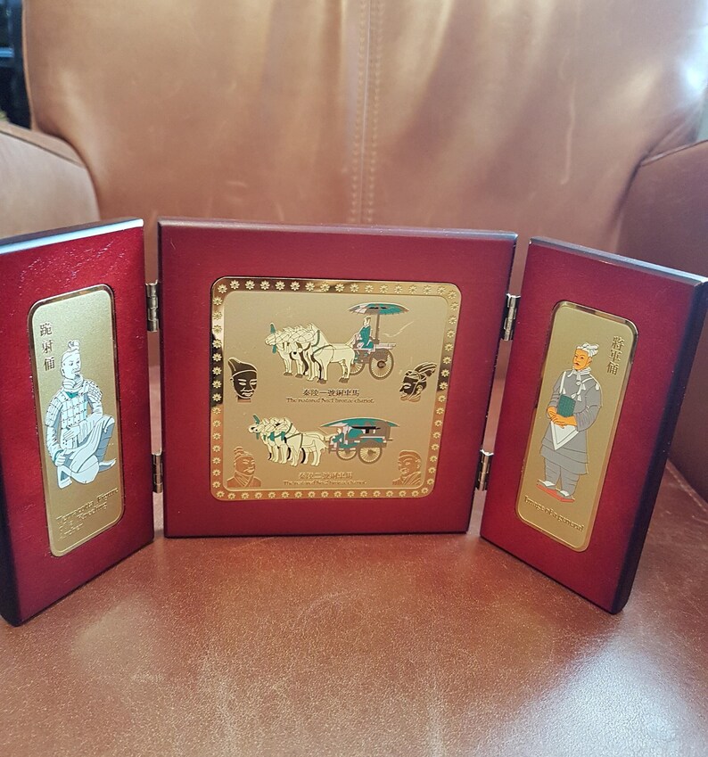 May include: A three-panel wooden display with gold-toned plaques depicting scenes from the Terracotta Army. The center panel shows a chariot pulled by four horses, while the side panels feature figures of warriors. The plaques are surrounded by a decorative border.