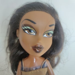 May include: A brown-skinned doll with long dark brown hair. The doll has green eyeshadow, brown eyeliner, and brown lipstick. The doll is wearing a brown and gold dress.