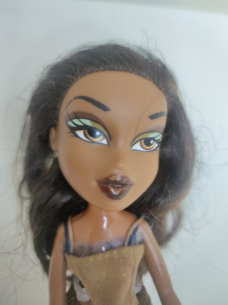 May include: A brown-skinned doll with long dark brown hair. The doll has green eyeshadow, brown eyeliner, and brown lipstick. The doll is wearing a brown and gold dress.