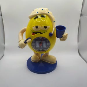 Può includere: Una sveglia a forma di personaggio M&M's giallo con un berretto da notte a pois blu. L'orologio ha una base blu e tiene uno spazzolino da denti e una piccola tazza blu. Il quadrante mostra il logo M&M's.