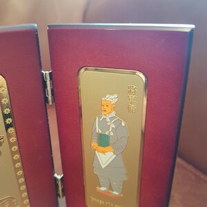 May include: A red wooden frame with a gold-colored inset featuring a portrait of a person in traditional clothing. The person is wearing a white hat and a gray robe. The text "將軍" is written in gold on the right side of the frame.