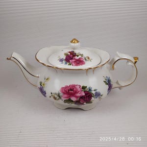 Op de afbeelding: Witte porseleinen theepot met een bloemmotief met roze en paarse rozen, geaccentueerd met gouden randen. Het deksel is versierd met een gouden knop en een bijpassend bloemmotief. Een klassiek item voor een theeservies.