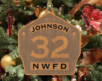 May include: A wooden Christmas ornament shaped like a fire station door plaque. It is personalized with the name "JOHNSON", the number "32", and the letters "NWFD". The ornament also includes the year "2025".